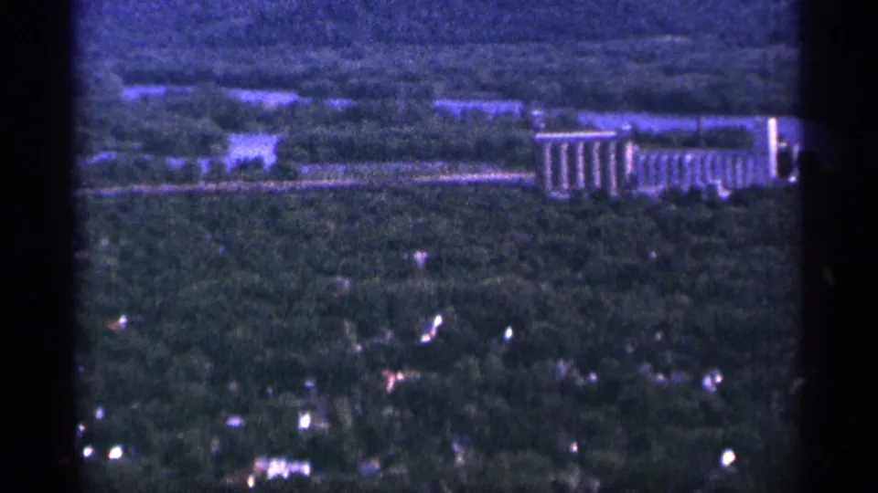 1965 Duluth aerial footage frame with wooded neighborhoods below and a long grain-elevator-like building near the horizon.