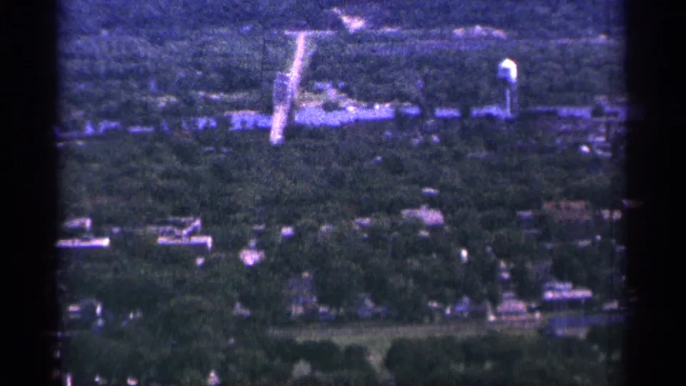 1965 Duluth aerial 8mm frame showing a road through trees, scattered homes, low buildings, and a water tower.