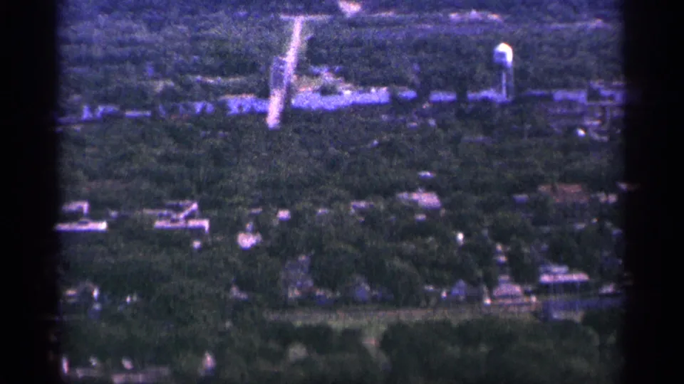 1965 Duluth aerial frame showing a water tower beside low buildings, with wooded residential streets in the foreground.