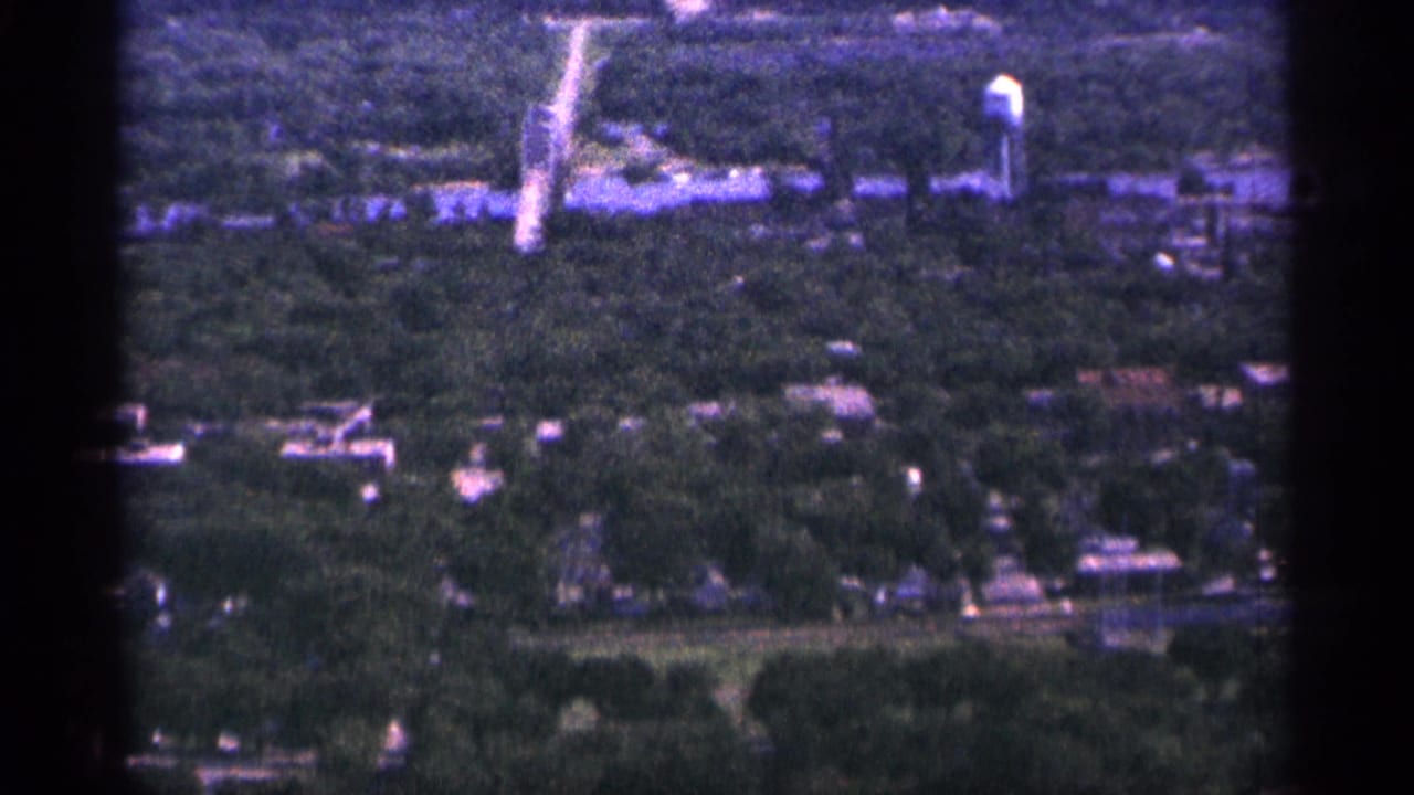 1965 Duluth home-movie frame showing a tree-filled townscape with scattered houses, roads, and a distant water tower.