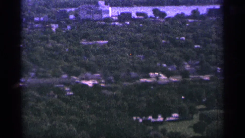 1965 Duluth home-movie frame with dense trees, scattered structures, roads, and a pale open area near the top edge.
