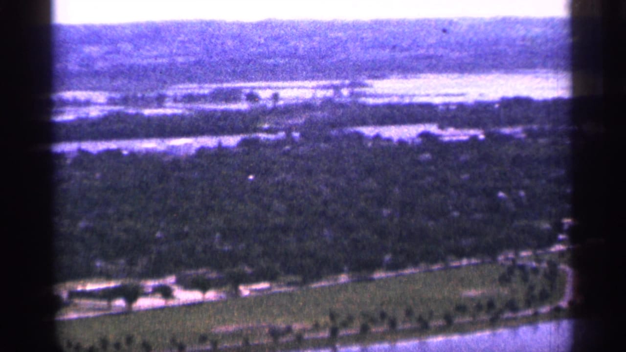 1965 Duluth aerial 8mm view showing wooded shoreline, broad water, a curving road, and open green land from a helicopter perspective.