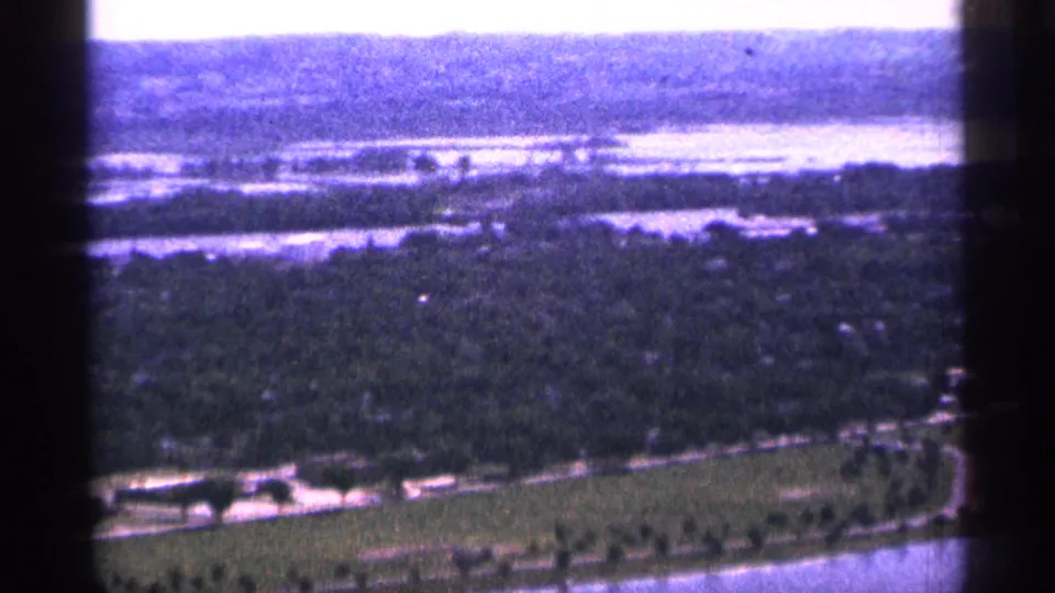 1965 Duluth aerial 8mm view showing wooded shoreline, broad water, a curving road, and open green land from a helicopter perspective.