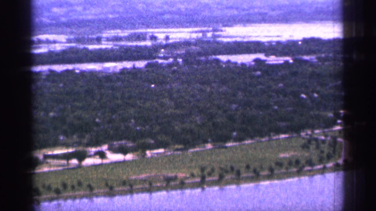1965 Duluth helicopter footage looking over tree-covered land, water channels, roads, and a distant shoreline under soft 8mm color shift.