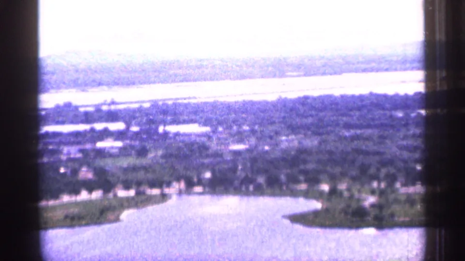 1965 Duluth aerial frame with a bright water surface, curved shoreline, tree-lined land, roads, and hazy distant terrain.