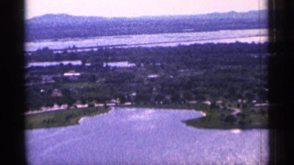 1965 Duluth aerial 8mm view of open water, a rounded shore edge, tree-lined roads, scattered buildings, and low distant hills.
