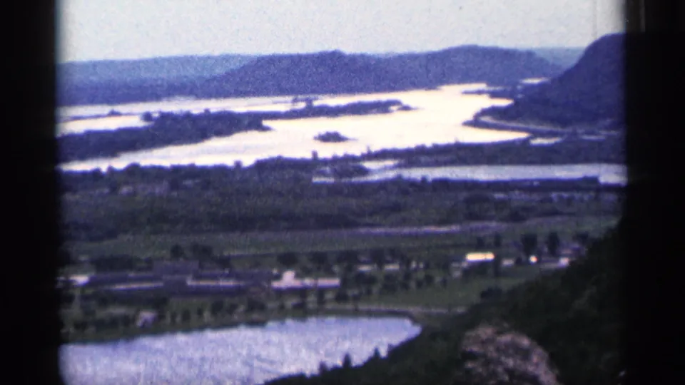 1965 home-movie frame from near Duluth showing broad waterways, green fields, rows of trees, and distant hills.