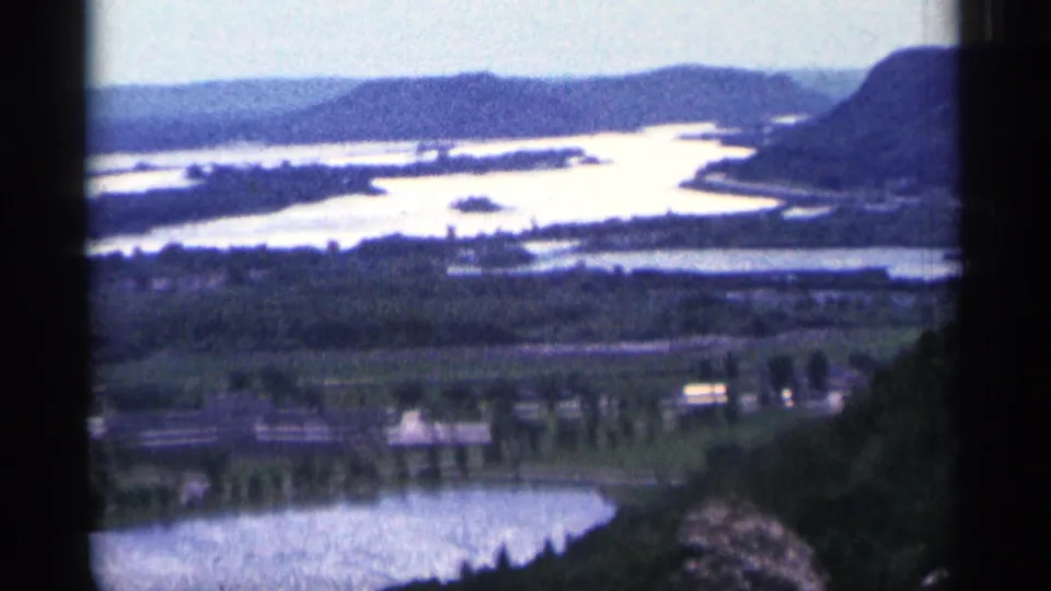 Duluth-area 1965 8mm view across lake edge, bright riverlike channels, wooded hills, and shoreline roads.