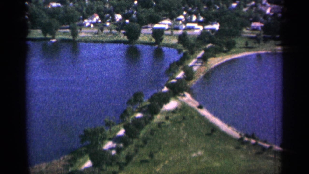 1965 Duluth 8mm aerial view over blue water, a curved shoreline, green lawns, trees, and small houses in the distance.