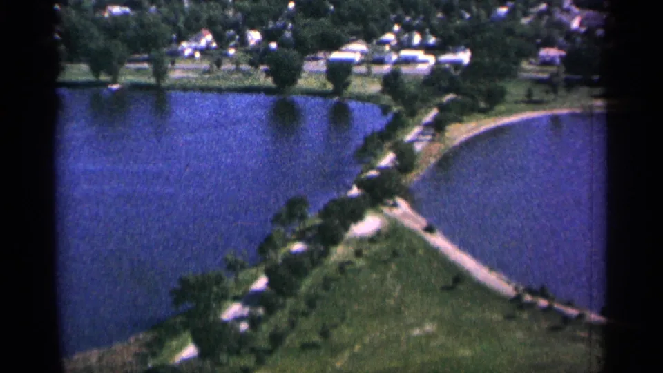 1965 Duluth 8mm aerial view over blue water, a curved shoreline, green lawns, trees, and small houses in the distance.