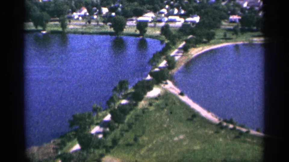 1965 Duluth aerial frame showing blue water on both sides of a narrow green area, with trees, paths, and distant homes.