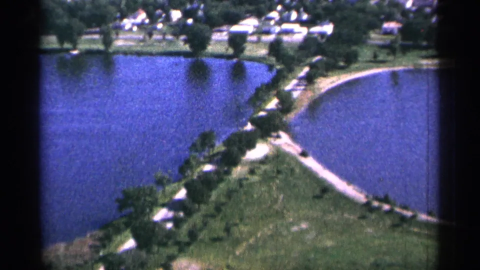 1965 Duluth 8mm aerial view looking across blue water toward a green shoreline with trees, paths, and small houses.