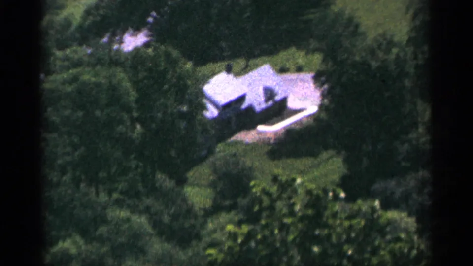 1965 Duluth aerial frame showing a white house set among dense trees, lawn, and surrounding foliage.