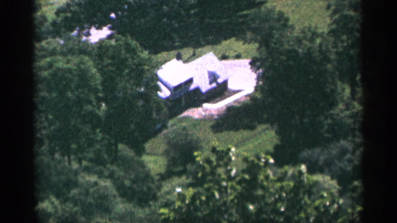 Distant white house surrounded by dense green trees and lawn.