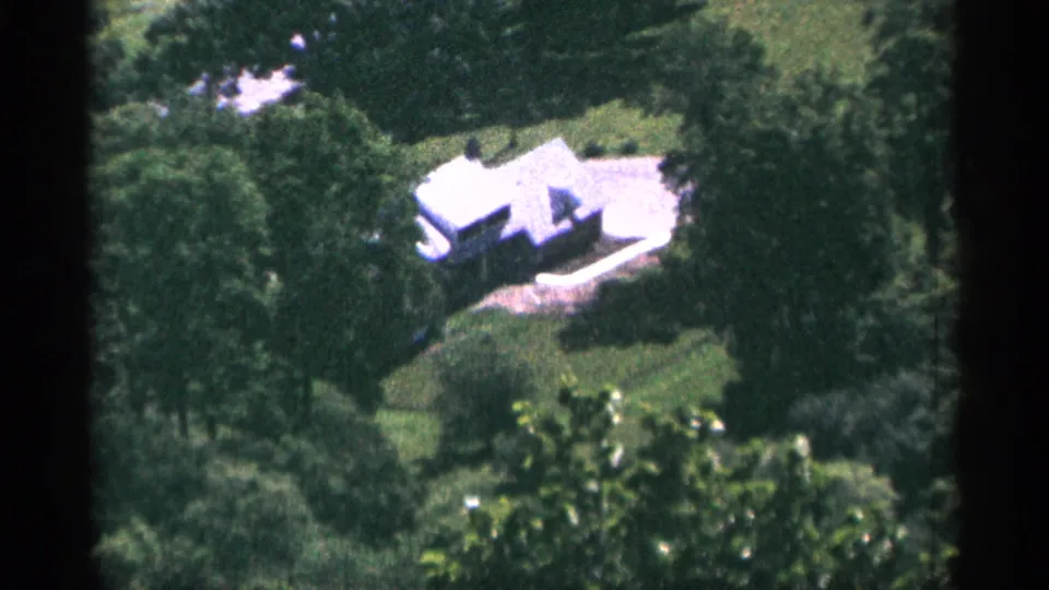 1965 Duluth home-movie frame showing a distant white house nestled among trees from an elevated viewpoint.