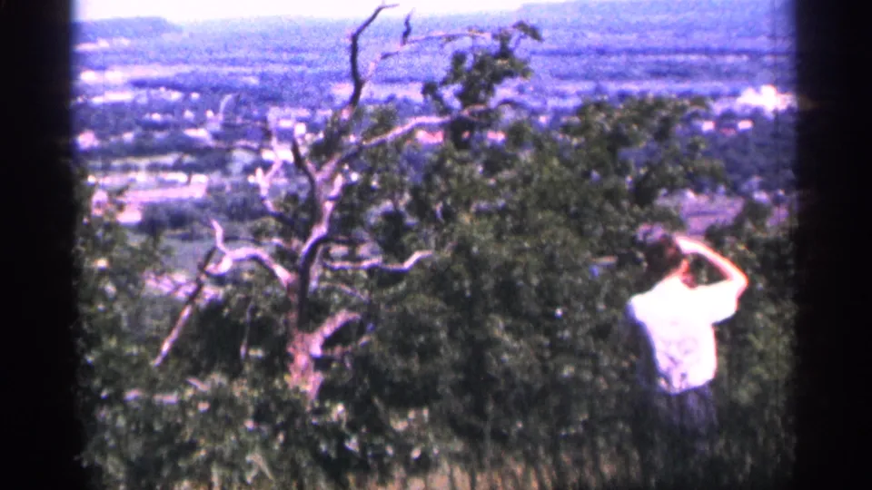 Adult man at a 1965 Duluth overlook appears to raise a camera or hand near his face, with trees and a distant town behind him.