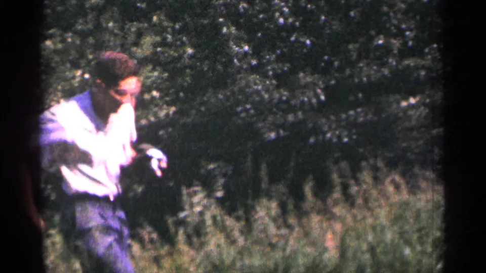 A man in a light shirt moves through the hillside grass, holding a small camera against a background of bushes and trees in the Duluth home movie.