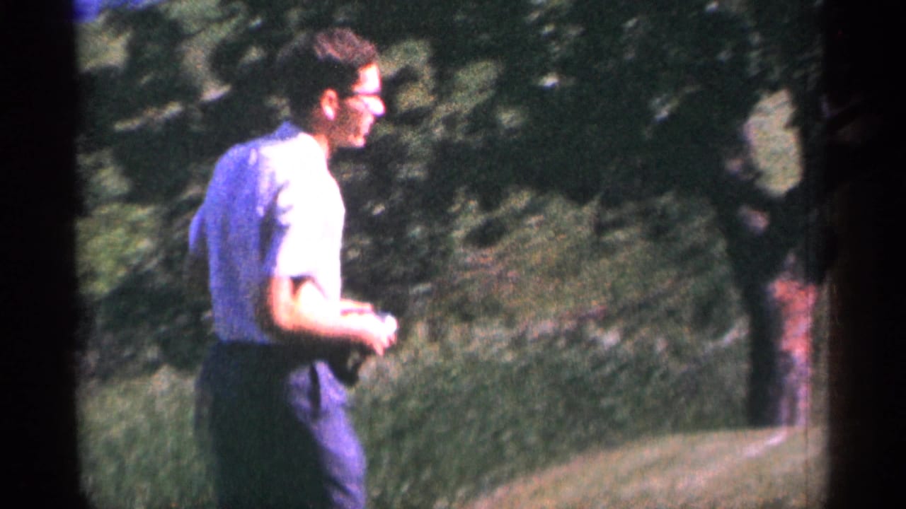 Man in glasses walks beside grass and trees in an outdoor area.