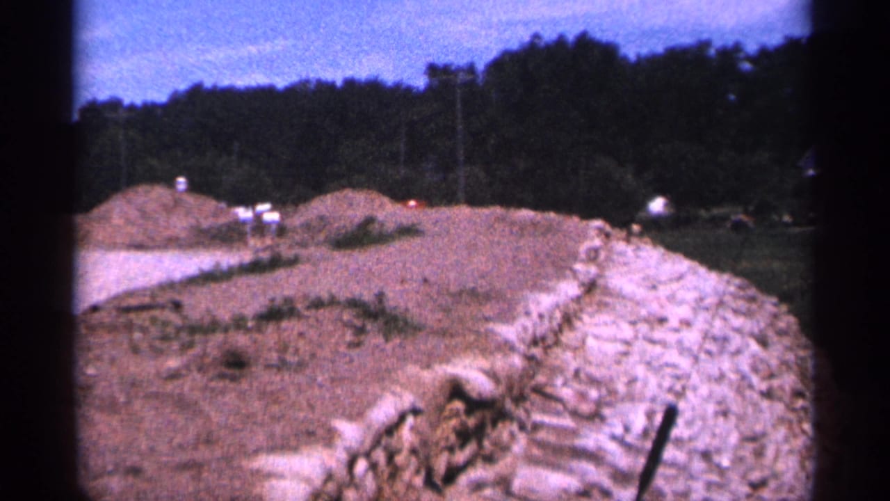 Dirt road construction area with earth piles, trees, and a stone retaining wall.