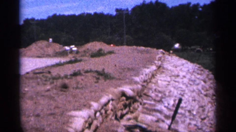 Duluth 1965 8mm frame showing a rough roadside area with a stone wall, dirt mounds, utility poles, and trees.
