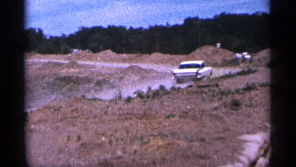 Duluth 1965 frame with a white 1960s sedan moving through a dusty unpaved roadside construction area.