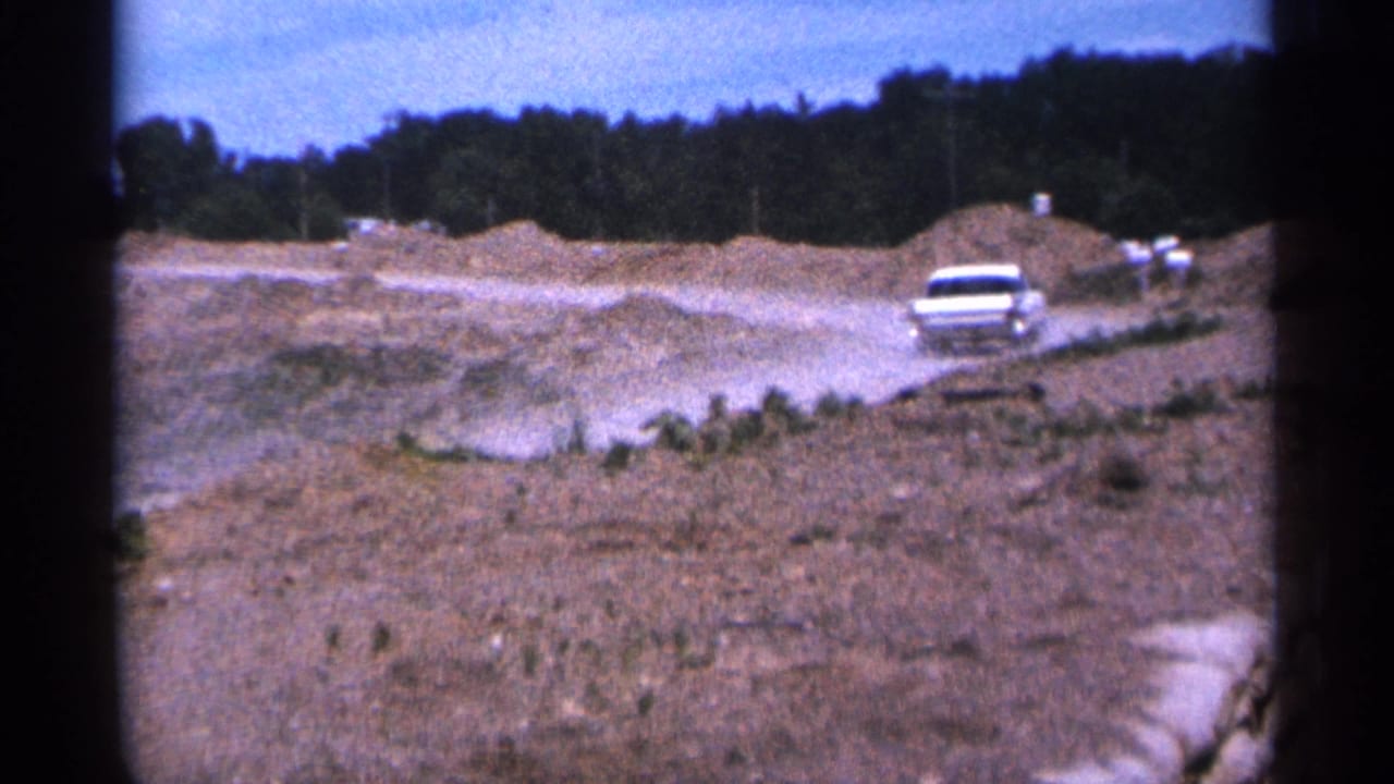 White car drives along a dusty dirt path beside earth piles and a treeline.