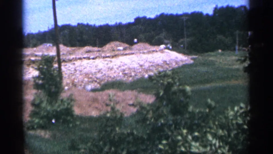 1965 Duluth home-movie frame showing grass, trees, utility poles, and a pale rock embankment under a blue sky.