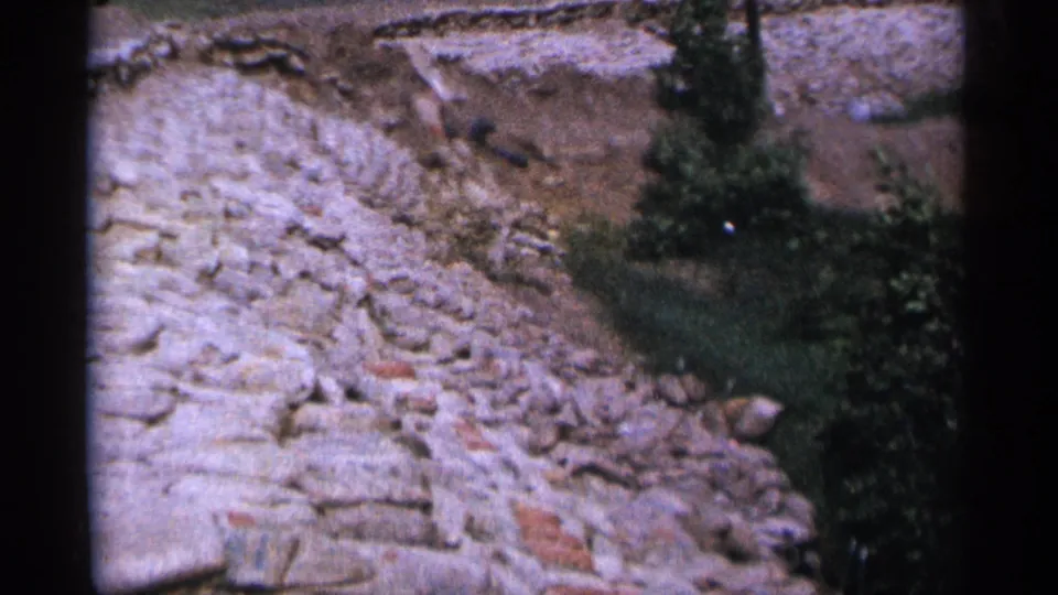 1965 Duluth home-movie frame showing a rock-and-dirt slope, grassy edge, shrubs, and a tree at the right side.
