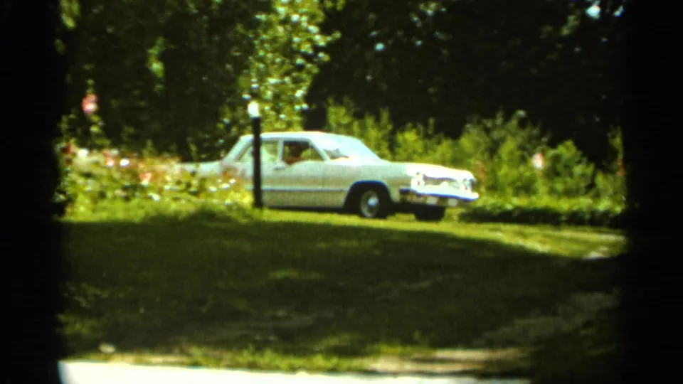 1965 Duluth home-movie frame showing a white sedan near a grassy driveway bordered by trees, shrubs, and a black lamp post.