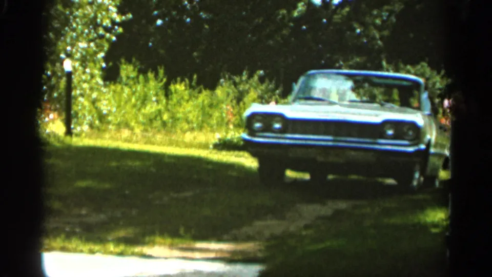 1965 Duluth home-movie frame showing the white sedan front-on along a grass-edged driveway, with trees and a lamp post in the background.