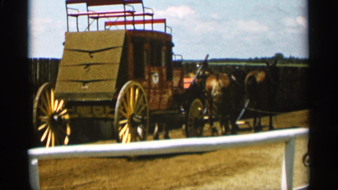 Horse-drawn stagecoach parked on a dirt lot behind a low barrier.