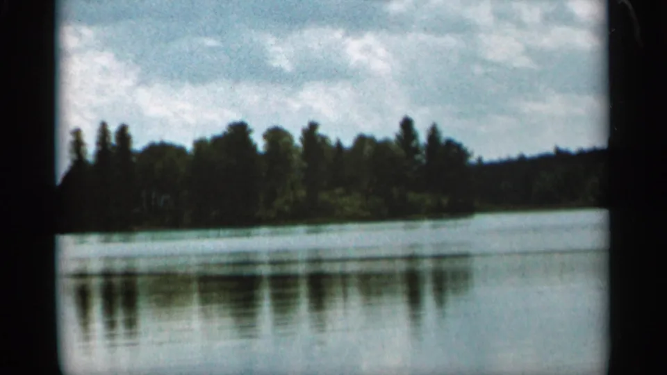 1965 Duluth-area 8mm frame showing a calm lake, dark tree-lined shoreline, and cloudy sky after the mule scene.
