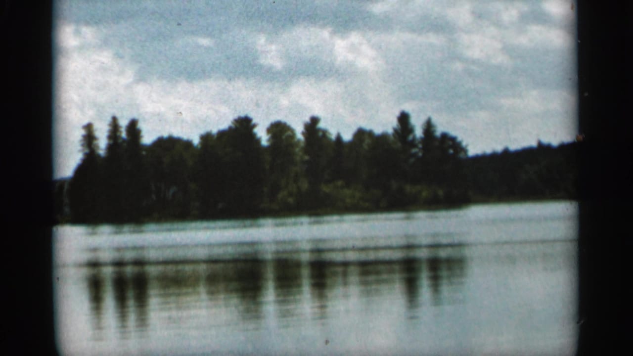 1965 Duluth-area 8mm frame showing a calm lake, wooded shoreline, and soft reflections beneath scattered clouds.