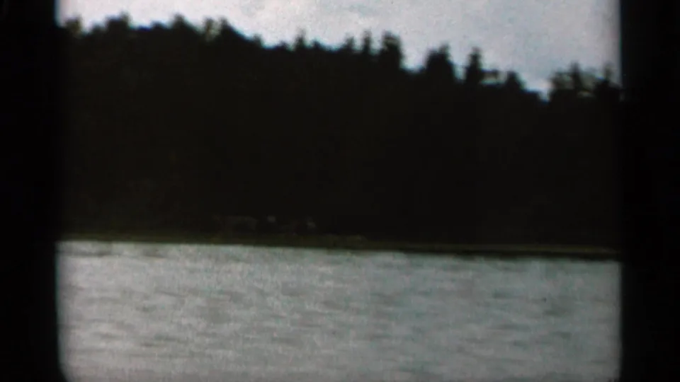 1965 Duluth-area waterfront view with lake water in the foreground, a dark tree line, and a small distant lodge-like structure.
