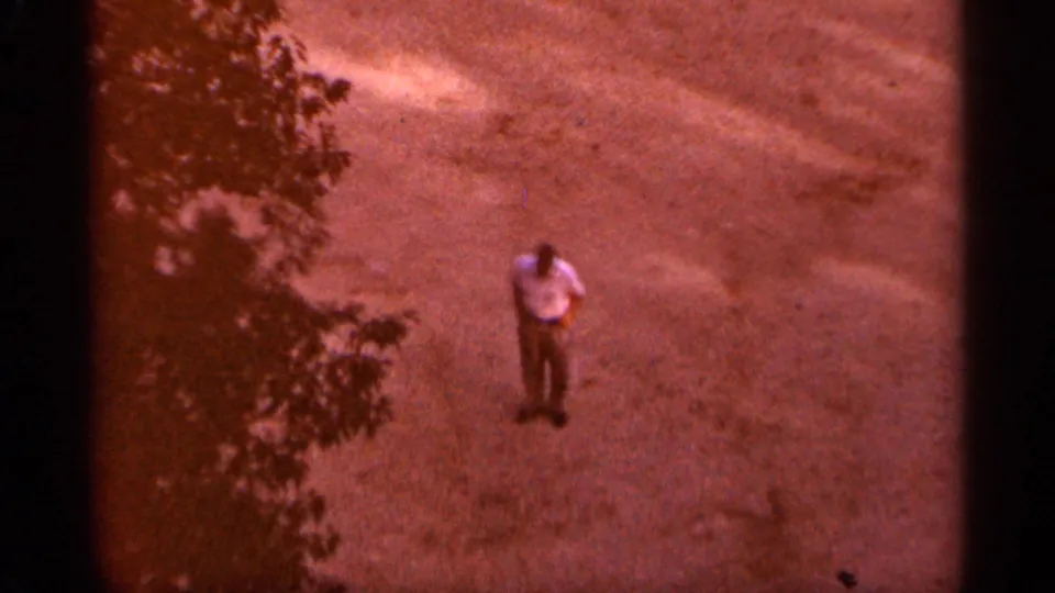 1965 Duluth home-movie frame with an adult man standing alone in an open gravel area near tree shade.