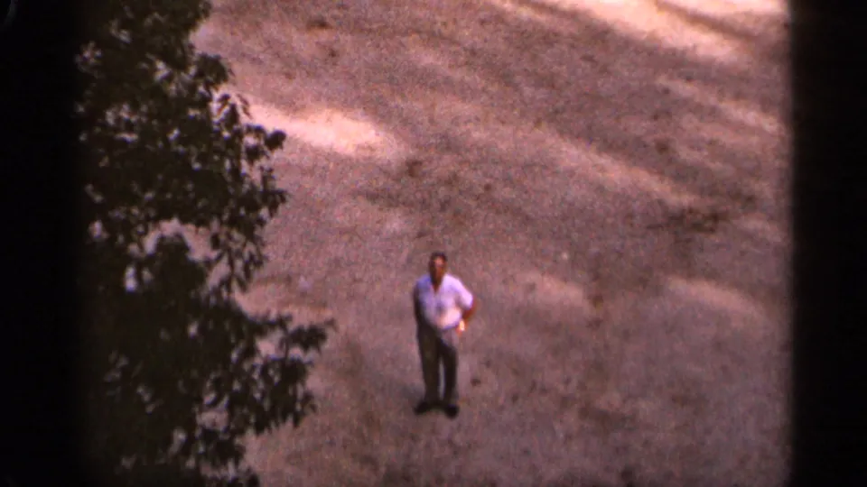 1965 Duluth home-movie frame showing an adult man in a light shirt standing on gravel near overhanging leaves.