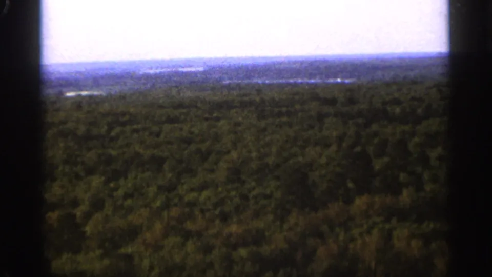 1965 Duluth-area 8mm frame looking over a broad forested landscape with faint distant water and a pale horizon.
