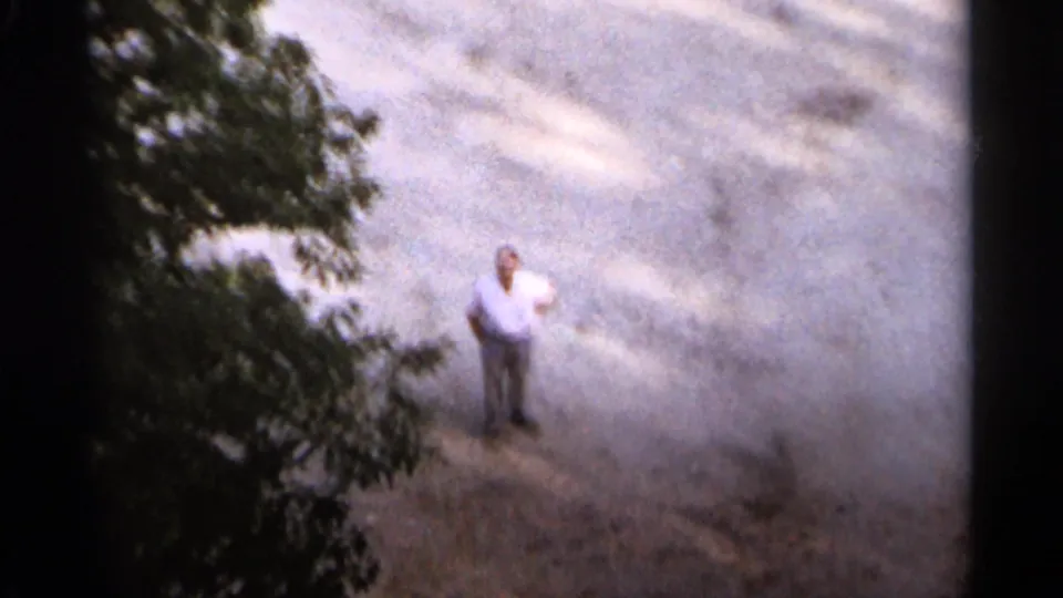 A man in a light shirt and dark pants stands on a pale clearing or road surface below the 1965 Duluth camera view.