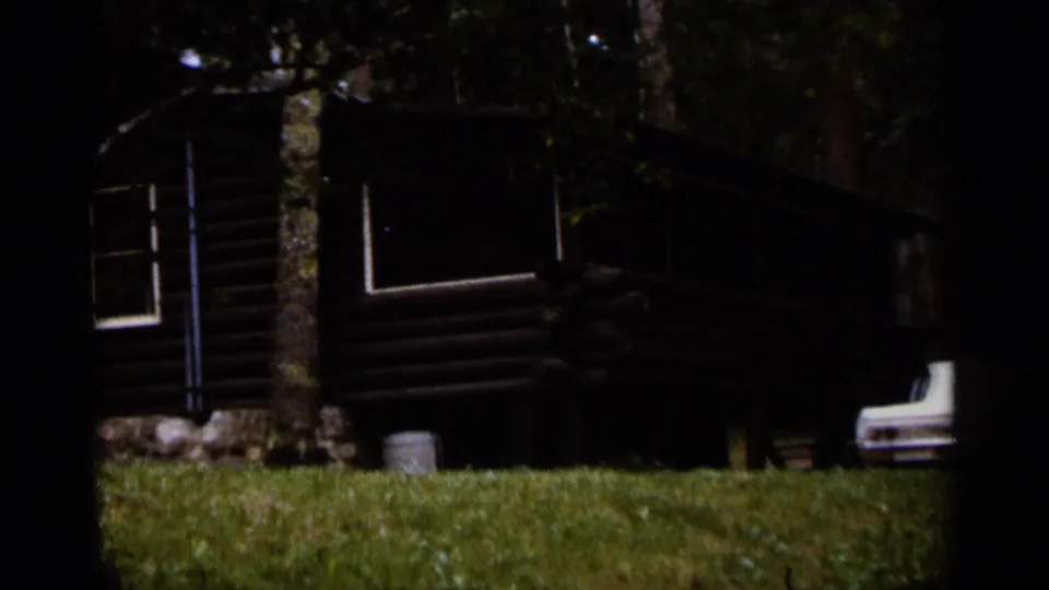 1965 Duluth home-movie view of a rustic log cabin in trees, grassy foreground, and a white car partly visible nearby.