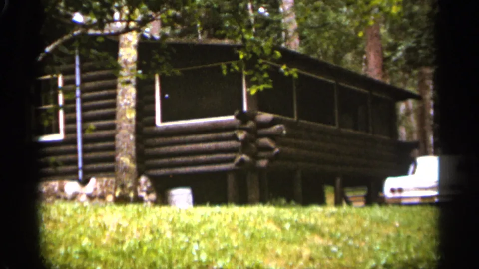 1965 Duluth frame showing a log cabin exterior, birch trees, grass, and a white period car in a wooded clearing.