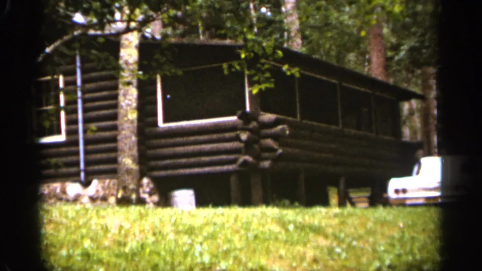 1965 Duluth-area home-movie frame showing a dark log cabin with a long screened porch, stone base, surrounding trees, grass, and a partially visible white car.
