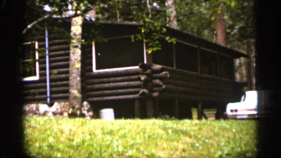 1965 Duluth-area frame of a rustic log cabin with a screened porch, dark siding, nearby tree trunks, grass, and the rear portion of a white car at the right edge.