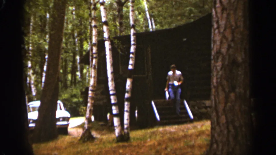 In the 1965 Duluth woods scene, a man exits the dark cabin carrying a white bin toward a vintage white car.