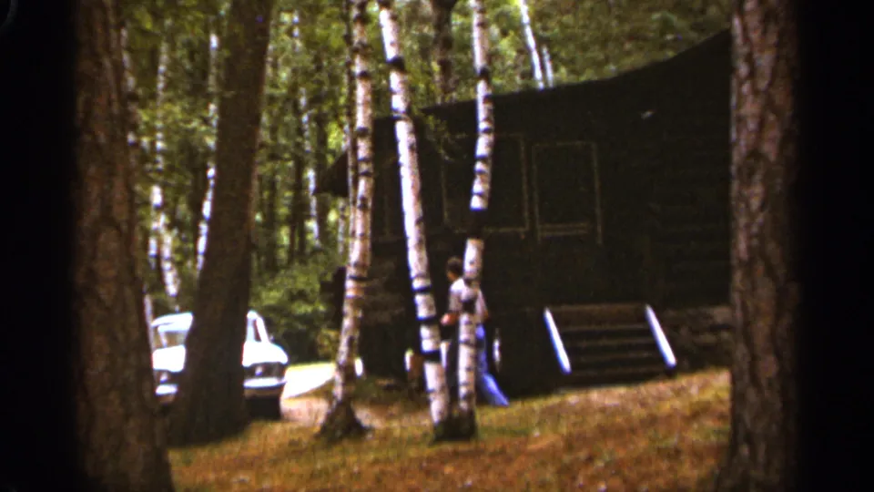 The man is near the cabin steps and birch trees, moving toward the parked white car in the Duluth 1965 home-movie scene.