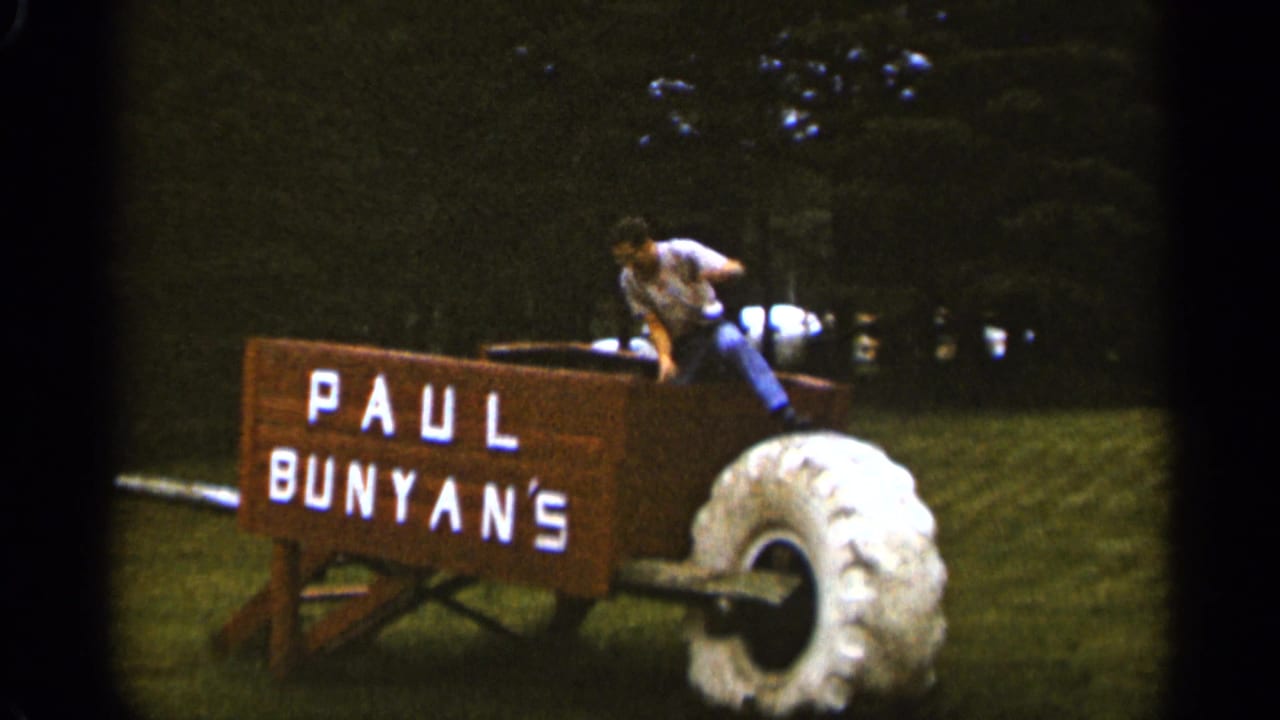 In 1965 Duluth, Minnesota footage, a man climbs into an oversized wooden wheelbarrow attraction with Paul Bunyan's painted on the side and a large white tire at right.