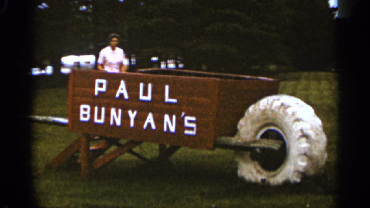 A woman stands inside the oversized Paul Bunyan's wooden wheelbarrow attraction on a grassy lawn in the supplied 1965 Duluth home-movie footage.