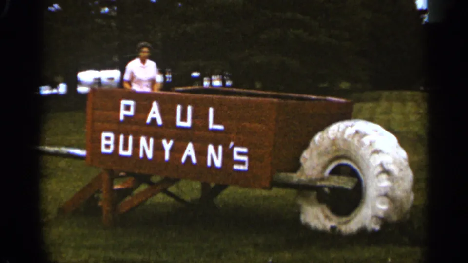 A woman stands inside the oversized Paul Bunyan's wooden wheelbarrow attraction on a grassy lawn in the supplied 1965 Duluth home-movie footage.