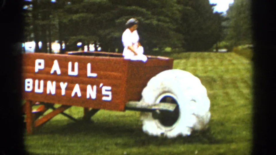A woman sits on the edge of the oversized Paul Bunyan's wheelbarrow attraction in the 1965 Duluth home-movie footage, with grass and trees behind her.