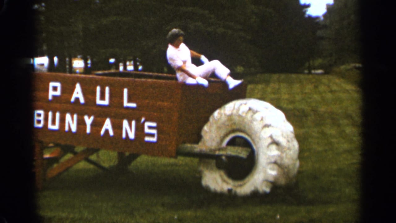In 1965 Duluth footage, a woman in white sits on an oversized wooden wheelbarrow marked Paul Bunyan's on a grassy lawn.