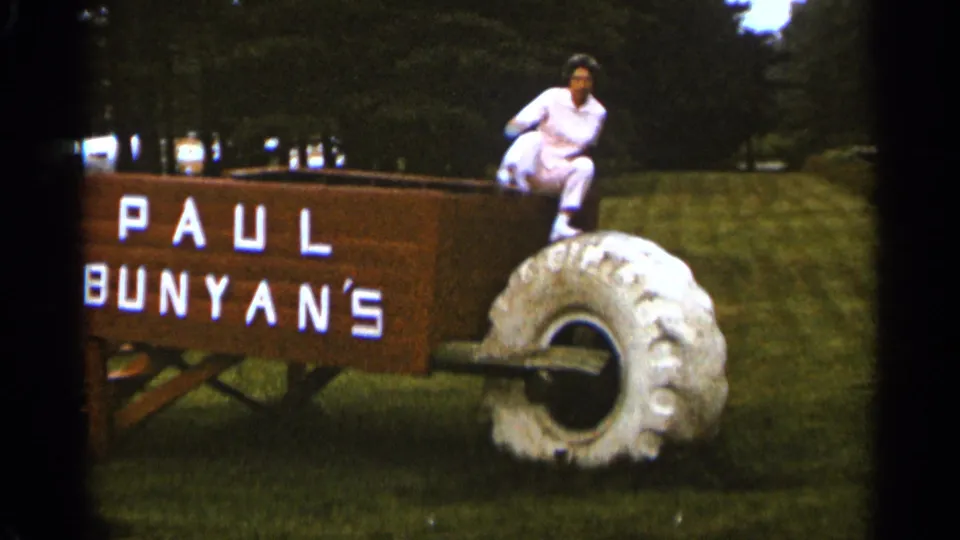 The woman appears to climb out of the Paul Bunyan's wheelbarrow display in a grassy Duluth setting, filmed on 8mm in 1965.
